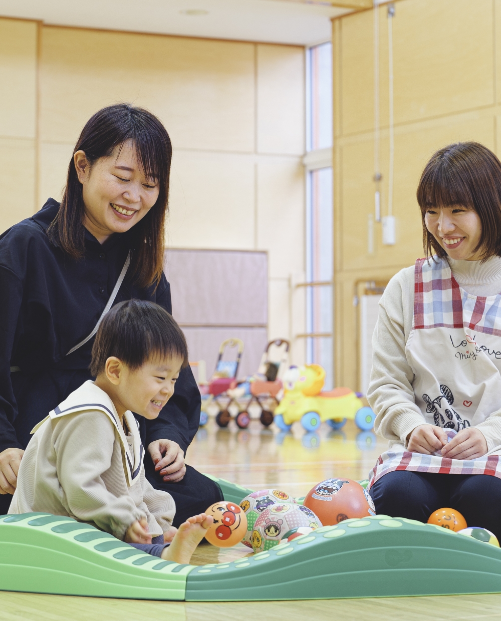 体育館のような部屋でボール遊びをする赤ちゃんと、それを見つめる保護者・児童館職員の写真